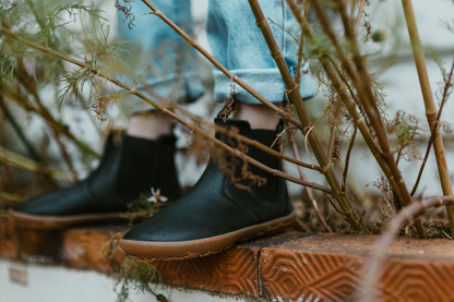 A person wearing PaperKrane black leather chelsea boots is standing on a ledge with some weedy looking plants around. 