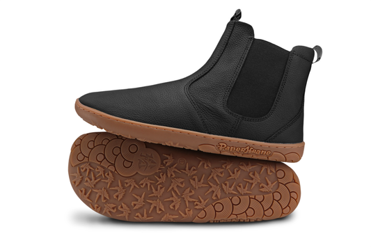 Black leather barefoot PaperKrane chelesa boots with gum soles on a white background. The view is of the side of one boot which sits on top of the other boot where you can see the foot-shaped sole and PaperKrane sole details. 