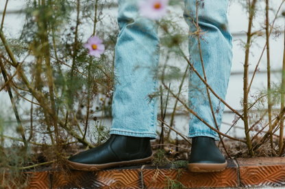 A person is standing in jeans (covering the top of the black leather PaperKrane chelsea boots) One foot is facing the camera and you can see the wider shape of the toe box. The person is surrounded by some plants. 