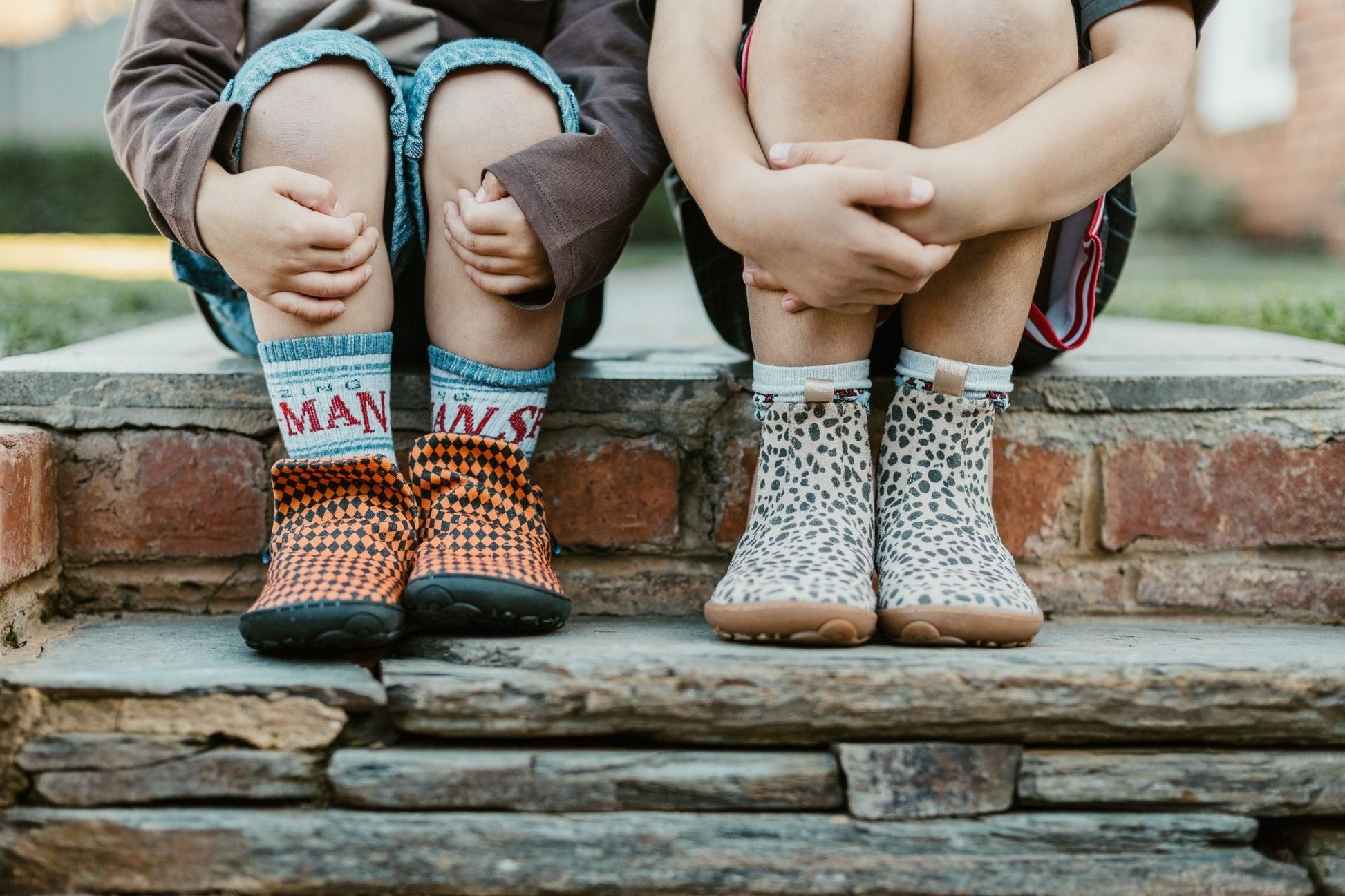 Two children sitting on stone steps outdoors, wearing two different PaperKrane barefoot models. The child on the left wears orange and black checkered signature style slip on shoes, and the child on the right wears beige and charcoal suede cheetah-print boots with white socks.