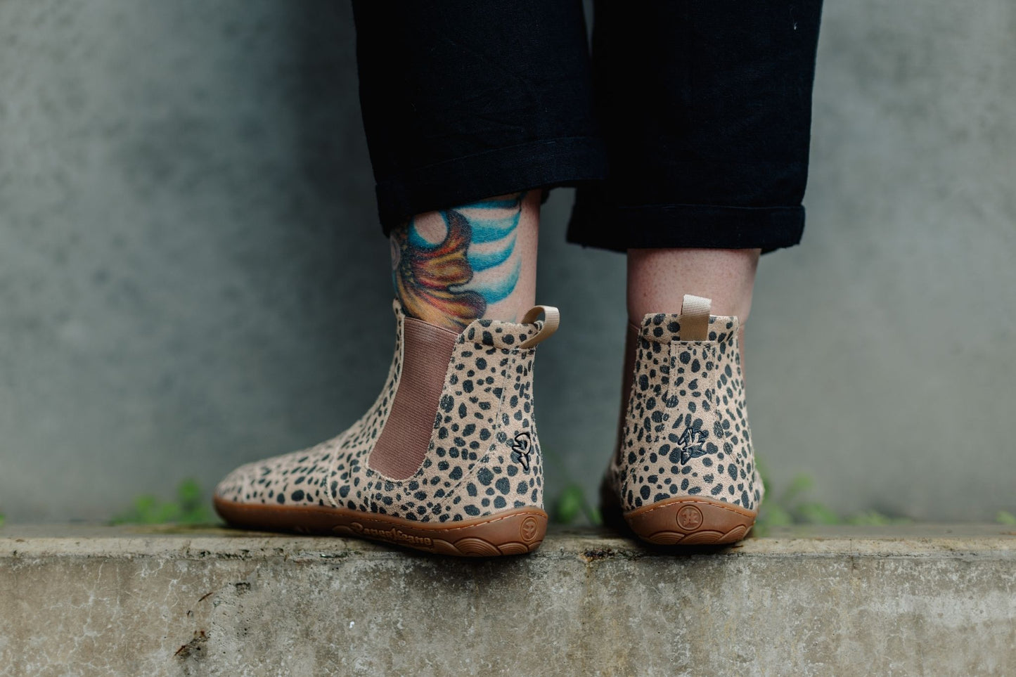 A person wearing PaperKrane cheetah-print chelsea boots with gum rubber soles, standing on a concrete ledge against a grey wall.