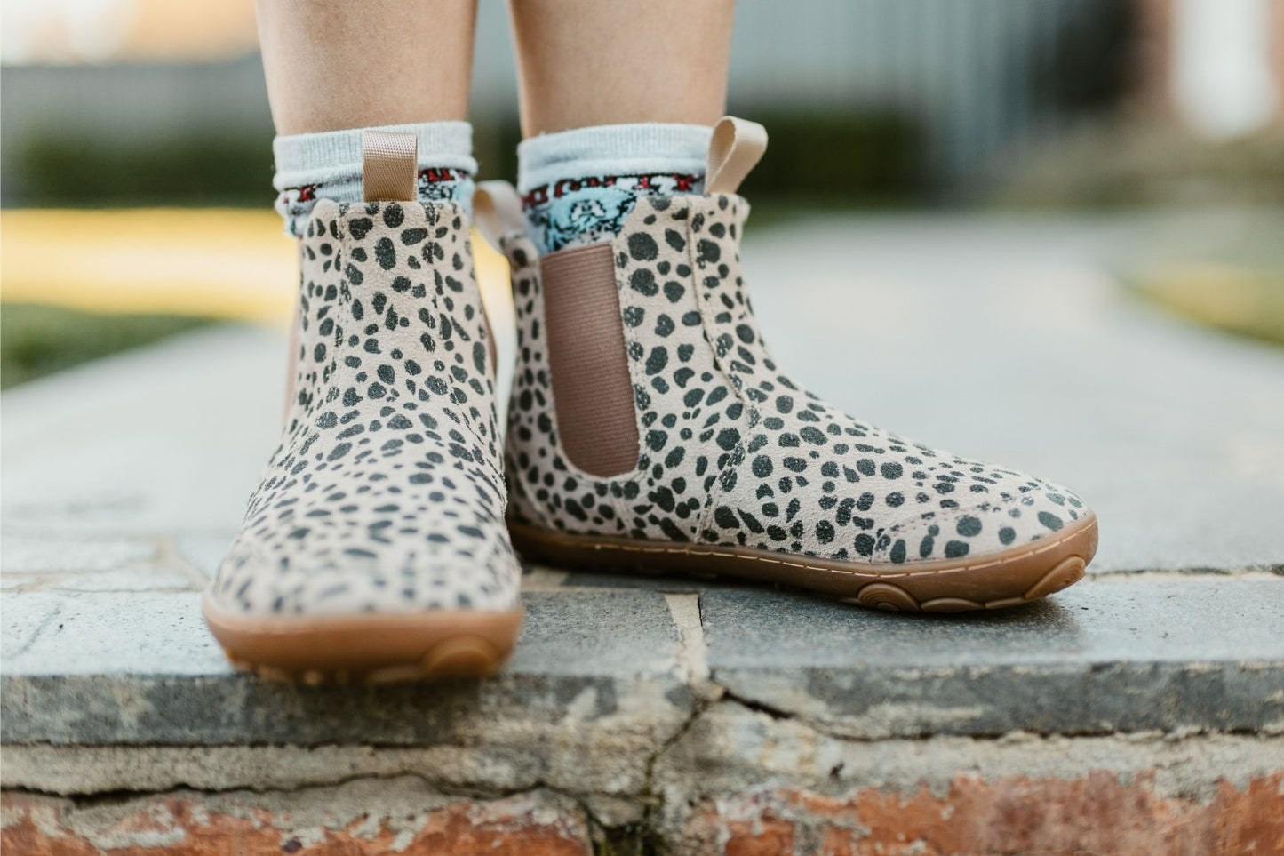 A child wearing beige and charcoal cheetah-print suede Chelsea boots with brown elastic side panels and gum soles, standing on a stone step outdoors, toes pointing forward.