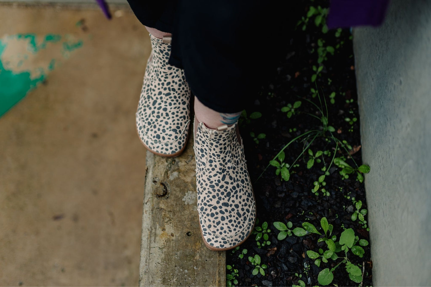 A person wearing suede cheetah-print foot-shaped chelsea boots and black pants, standing on a concrete ledge.