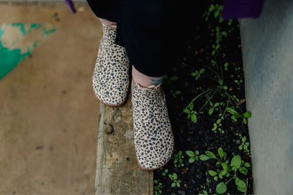 A person wearing suede cheetah-print foot-shaped chelsea boots and black pants, standing on a concrete ledge.