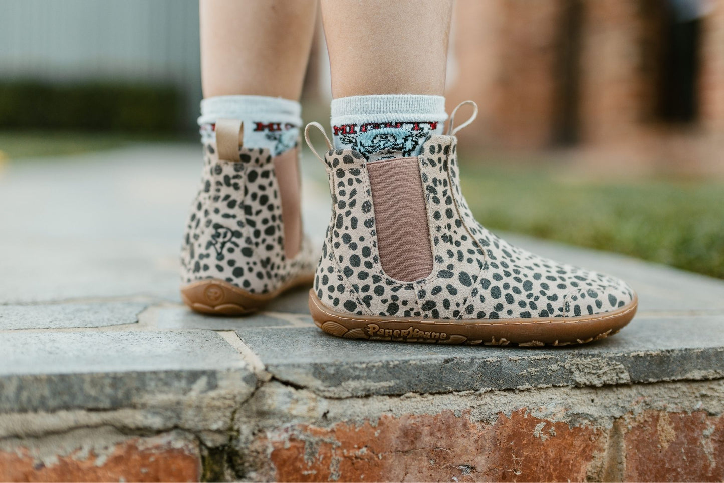 A child wearing beige and charcoal cheetah-print suede Chelsea boots with brown elastic side panels and gum soles, standing on a stone step outdoors.