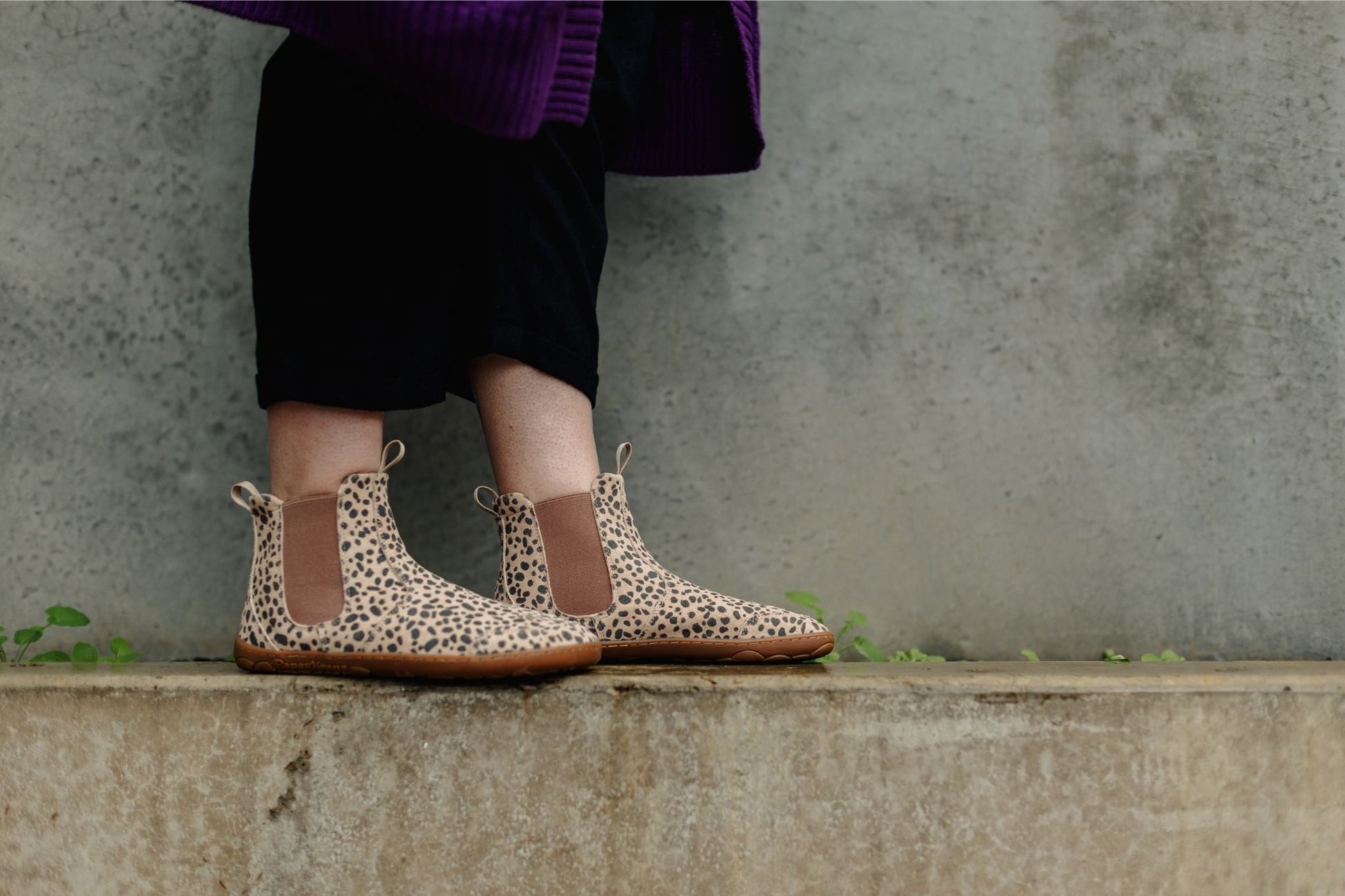 A person wearing PaperKrane cheetah-print Chelsea-style ankle boots with brown elastic side panels, standing on a concrete ledge against a textured grey wall.