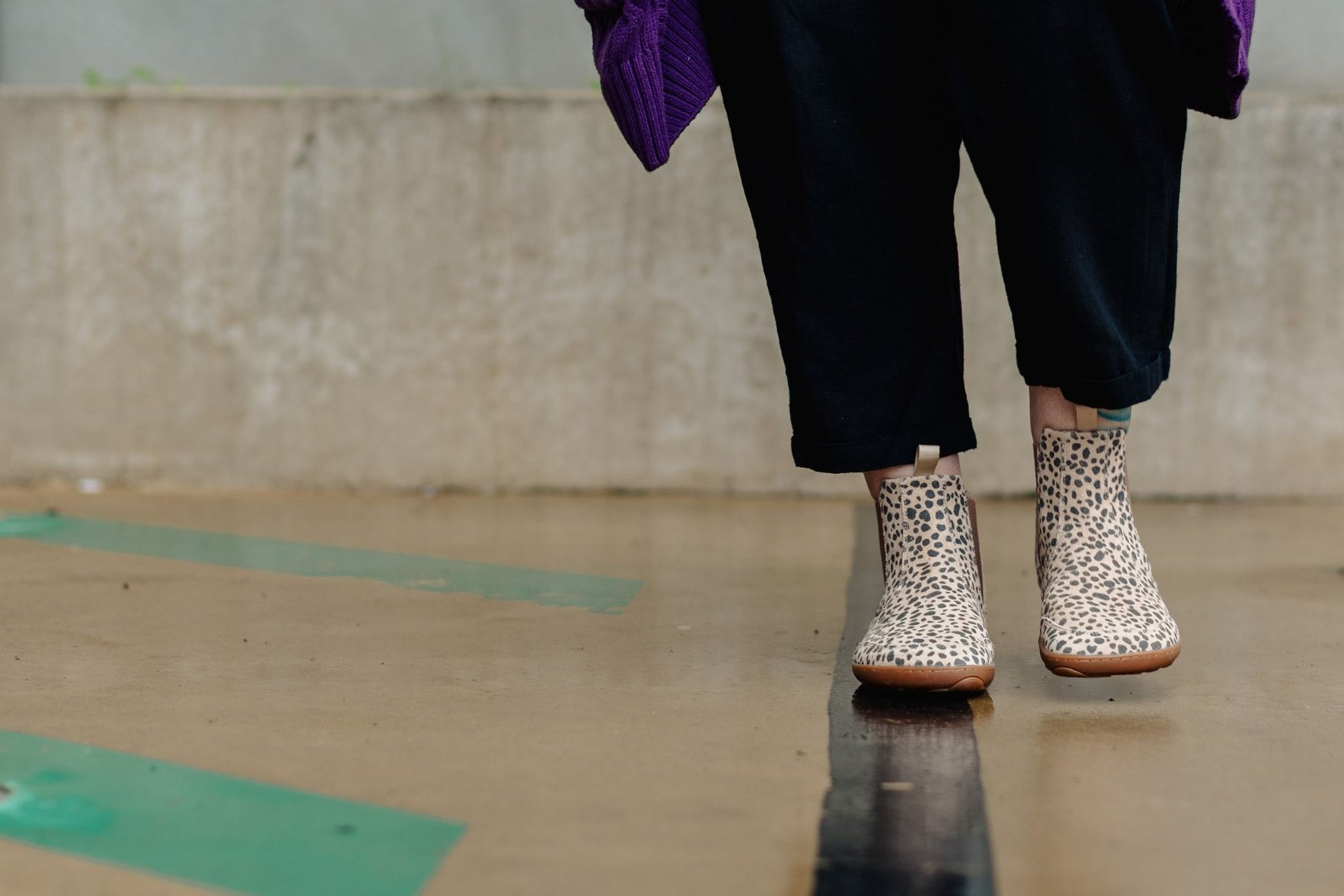 A person wearing beige and black cheetah-print barefoot chelsea boots with black pants, walking on a wet concrete surface.