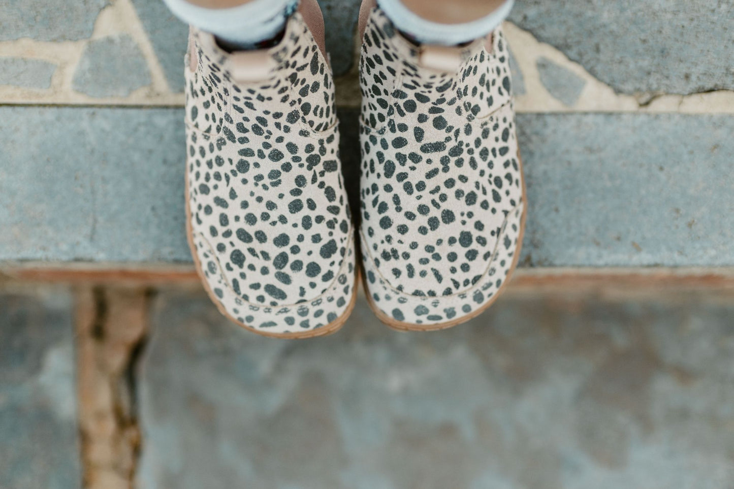 A child wearing beige and charcoal cheetah-print suede Chelsea boots with brown elastic side panels and gum soles, standing on a stone step outdoors, the view is from above the foot-shaped toe box.