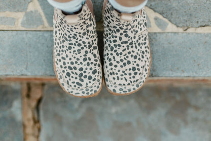 A child wearing beige and charcoal cheetah-print suede Chelsea boots with brown elastic side panels and gum soles, standing on a stone step outdoors, the view is from above the foot-shaped toe box.