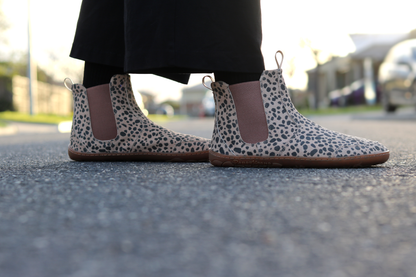 Side view of cheetah print suede chelsea boots by PaperKrane. The person wears dark socks and pants, and the background is on a street. 