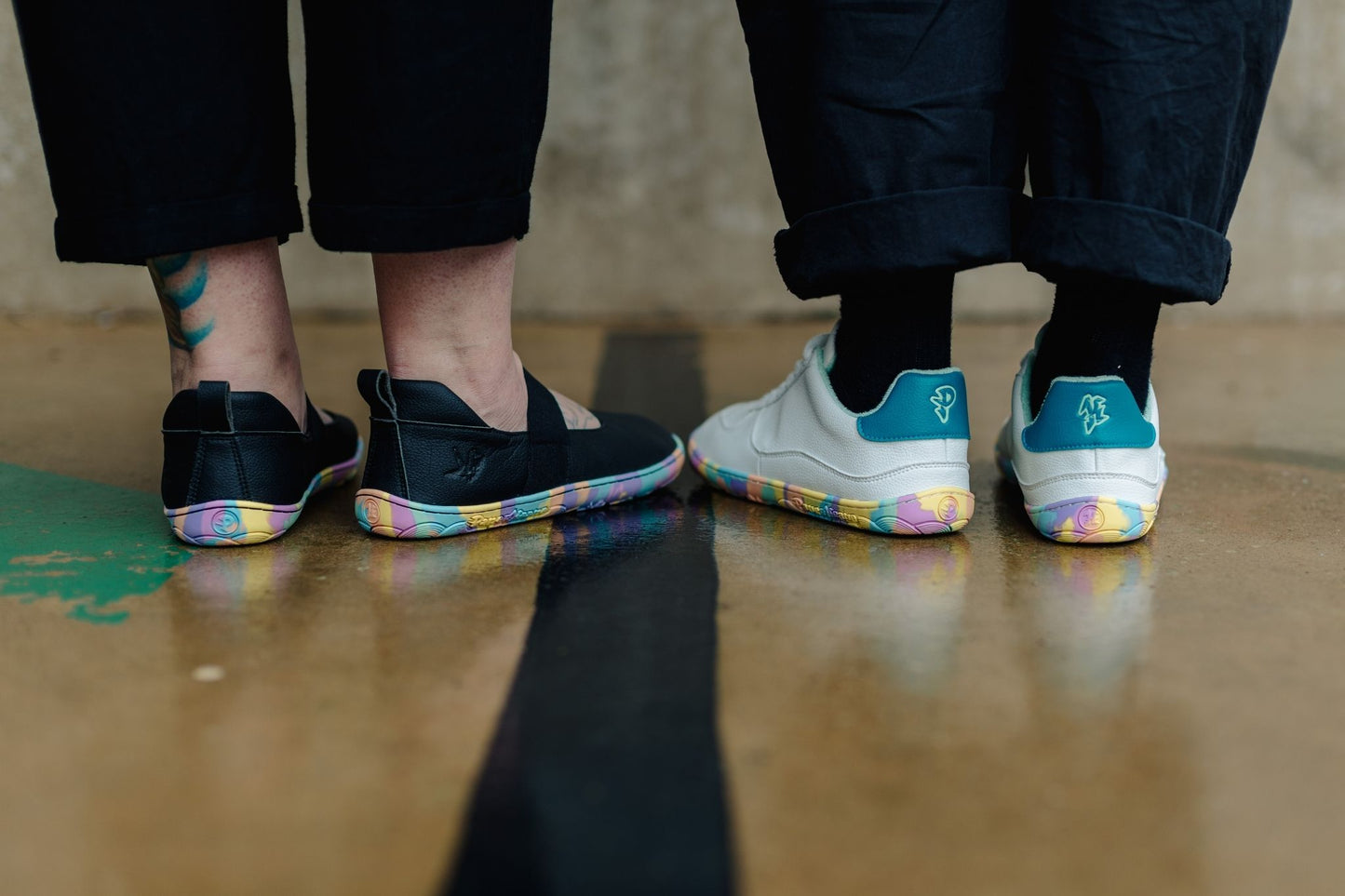 Two people standing side by side, shown from the calves down, wearing colourful-soled mjs and sneakers. The person on the left wears black slip-on mary janes, and the person on the right wears white sneakers with teal heel accents and rainbow-coloured soles.