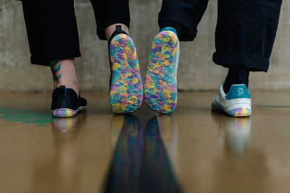 Two pairs of shoes shown from the bottom, featuring a black slip-on mj, and a pair of white vegan leather sneakers with colourful rainbow-patterned soles, set against a concrete wall and floor.