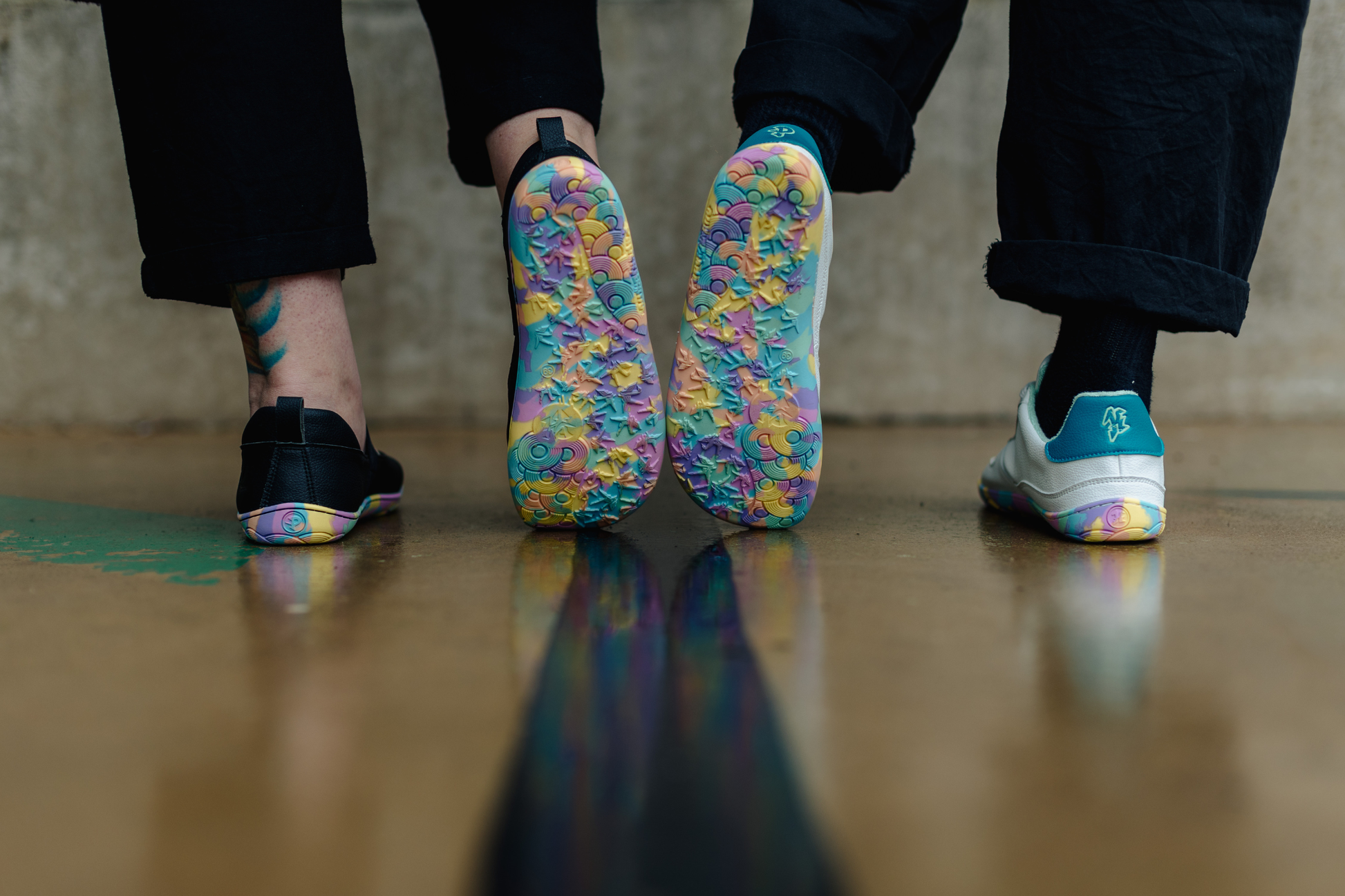 Two pairs of shoes shown from the bottom, with the centre two feet turned to display a highly textured, multicoloured patterned rubber soles. The shoes are black and white mjs and sneakers, while the right shoe is white and teal, the left are all black uppers with elastic strap over the foot.