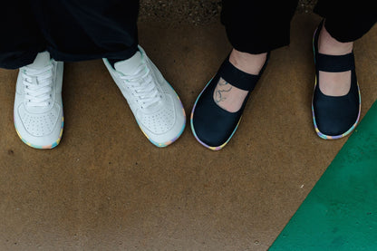 Two people standing on a brown concrete surface, shown from the knees down. One person wears white sneakers with rainbow-coloured outsoles, and the other wears black Mary Jane-style shoes with matching rainbow outsoles.