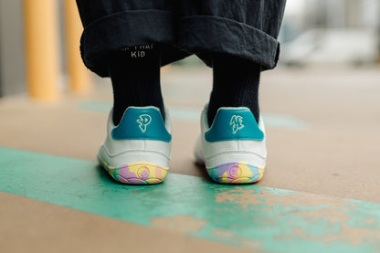 White vegan leather PaperKrane barefoot sneakers with teal heel accent and multi-coloured rainbow rubber soles, viewed from the back on a concrete surface. 