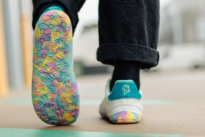 A close-up lifestyle shot of a person's feet wearing colourful, rainbow-patterned sneakers. The left shoe is shown from the sole, revealing a vibrant, multicoloured abstract design in shades of yellow, green, blue, and pink.