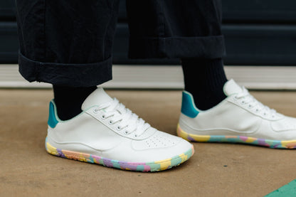 White low-top barefoot sneakers with teal heel accents and multicolour rainbow detailing on the outsole, shown on a person wearing black pants and socks.