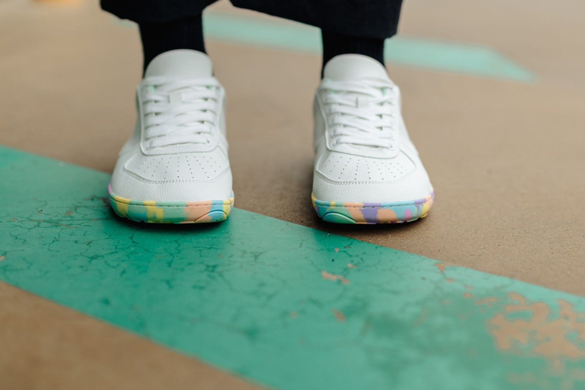A close-up shot of a person's feet wearing white low-top foot-shaped sneakers with colourful rainbow-patterned soles, standing on a green painted concrete surface.