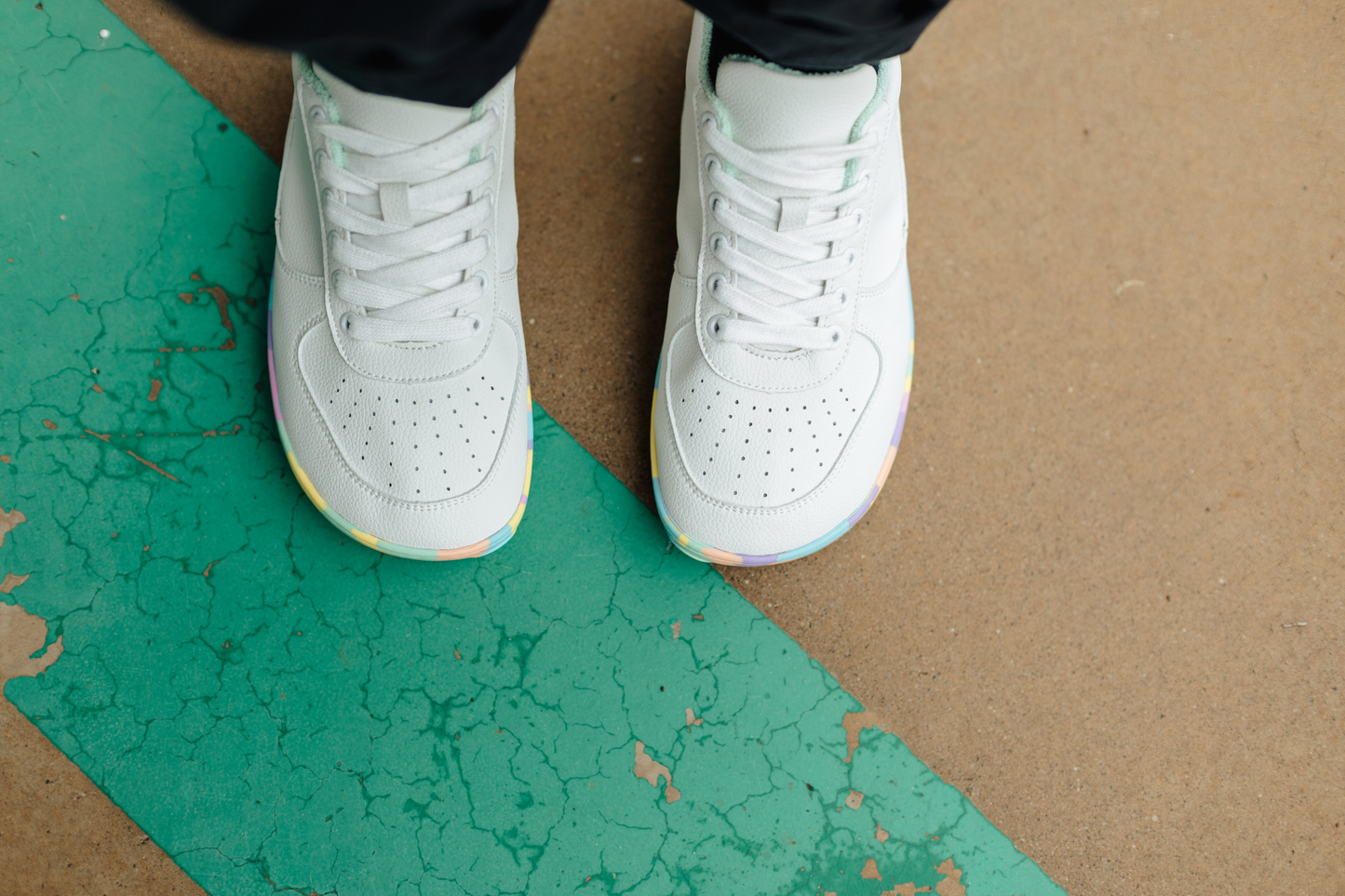A top-down view of a person wearing white foot-shaped PaperKrane sneakers with rainbow-coloured soles, standing on a brown concrete floor with a cracked teal painted stripe.