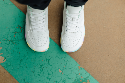 A top-down view of a person wearing white foot-shaped PaperKrane sneakers with rainbow-coloured soles, standing on a brown concrete floor with a cracked teal painted stripe.