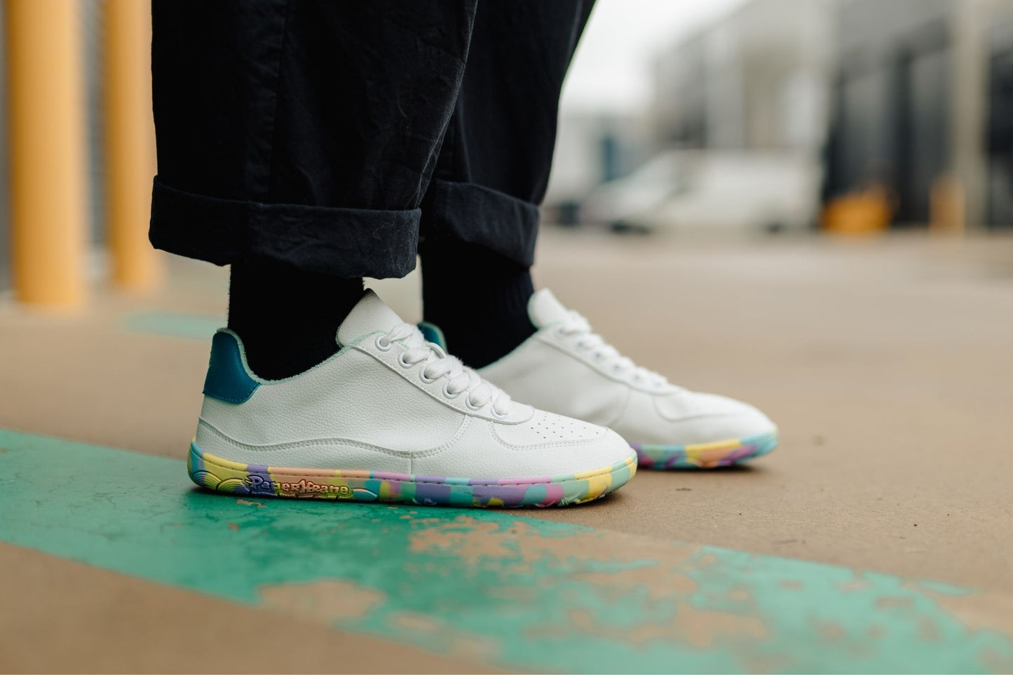 White low-top barefoot PaperKrane sneakers with colourful rainbow soles and teal heel accents, worn with black pants and socks on an outdoor concrete surface.
