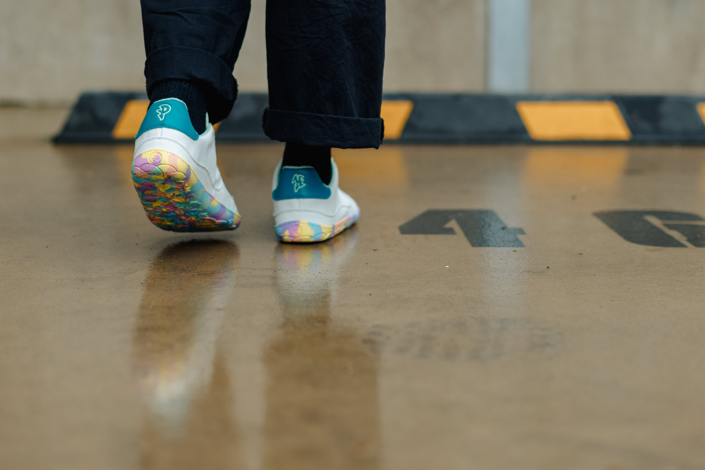 A person wearing white sneakers with colorful rainbow-patterned soles and teal heel accents, walking on a wet concrete floor with a black and yellow speed bump in the background.