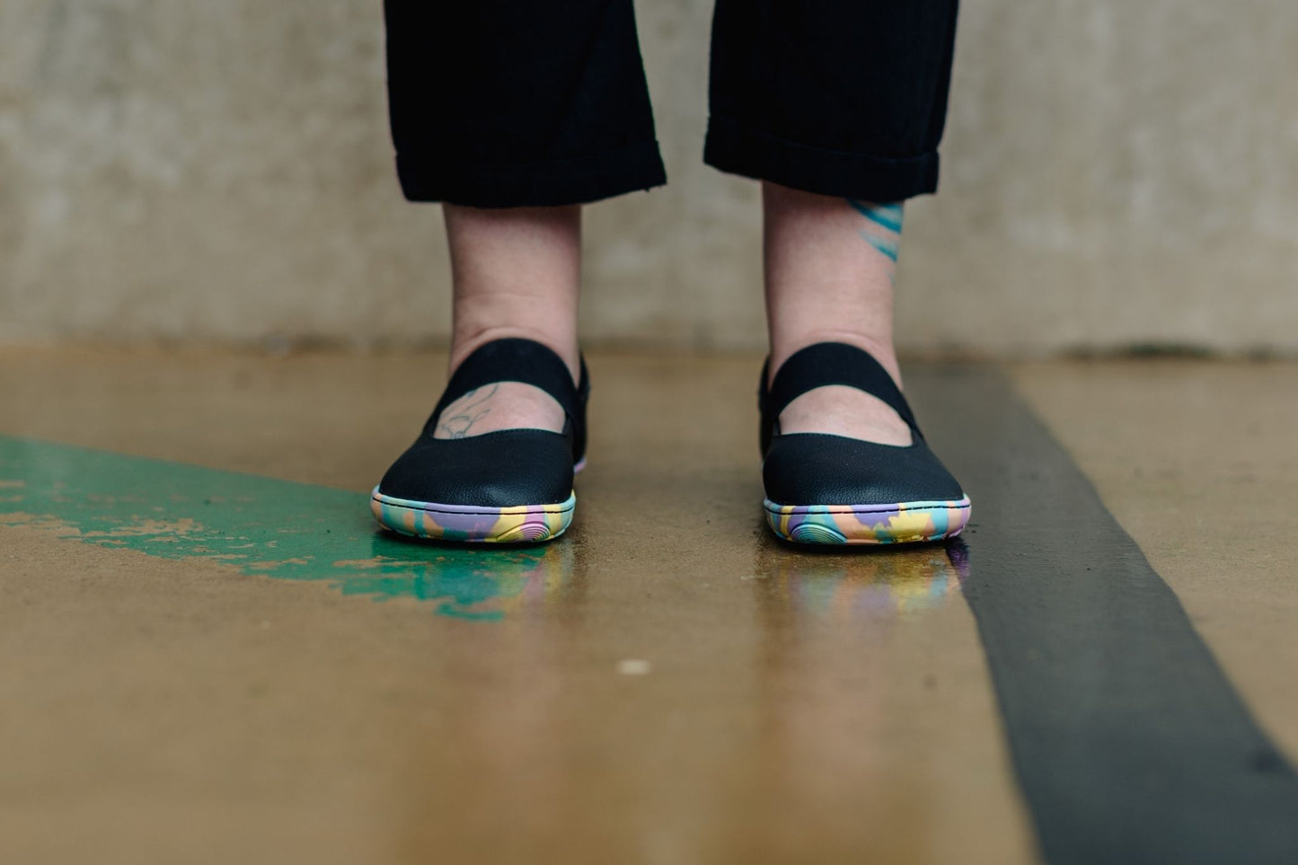 A person wearing black Mary Jane flats with colourful rainbow patterned soles, standing on a wet concrete floor.