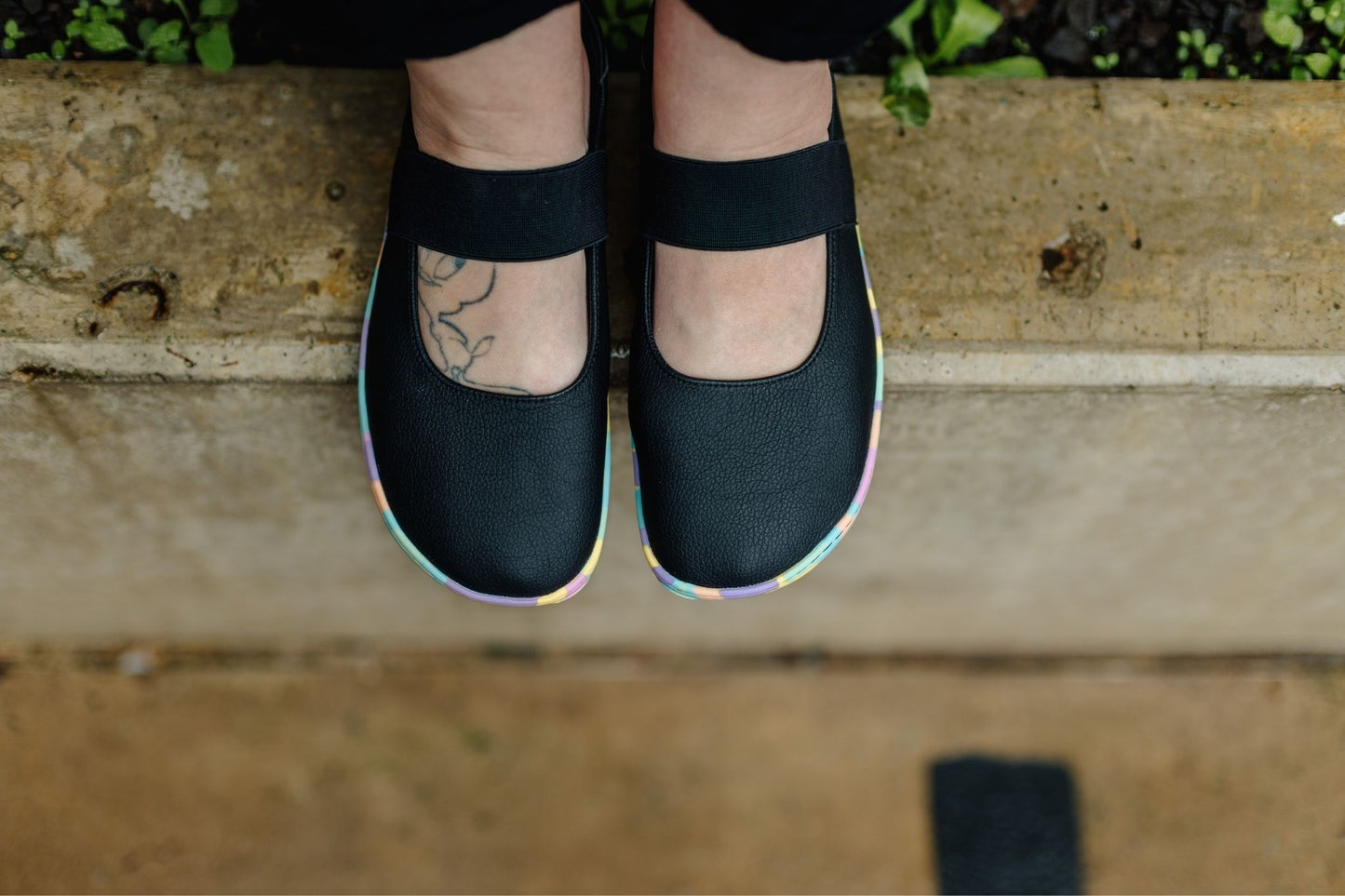 Black Mary Jane style shoes with splatter style bright pastel rainbow soles, shown on a person's feet from above, standing on a concrete ledge.