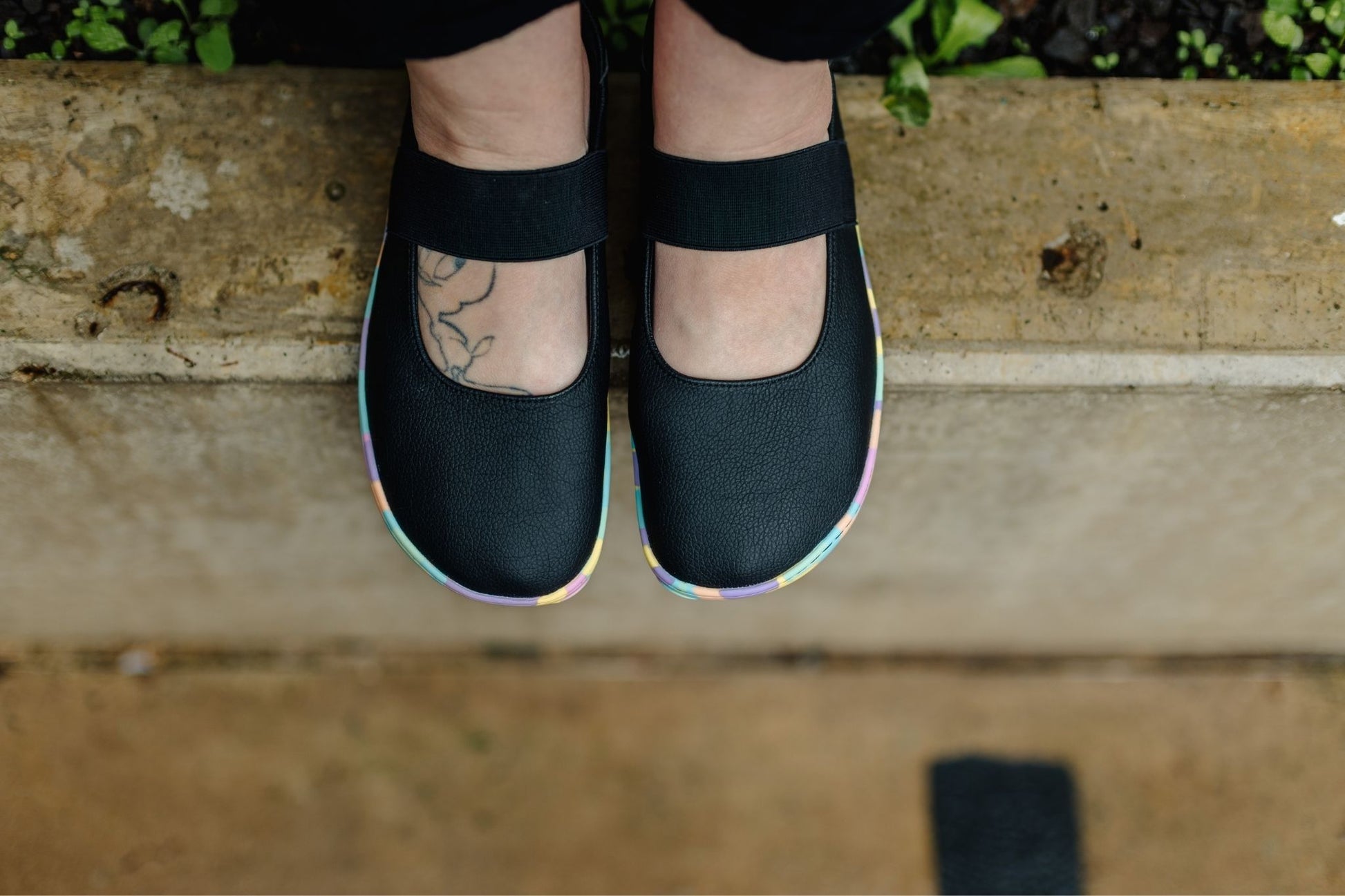 Black Mary Jane style shoes with splatter style bright pastel rainbow soles, shown on a person's feet from above, standing on a concrete ledge.