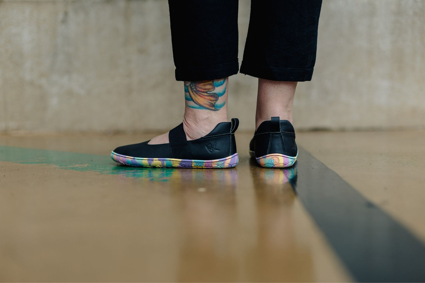 A person wearing black vegan leather PaperKRane mary janes with colourful rainbow-patterned soles, standing on a wet concrete surface in front of a concrete wall.