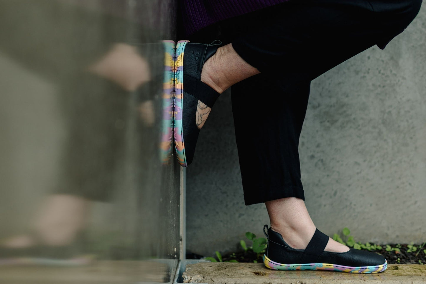 A close-up lifestyle shot of a person wearing black slip-on PaperKrane mary janes with a colourful, rainbow-like sole and black pants, sitting against a concrete wall.