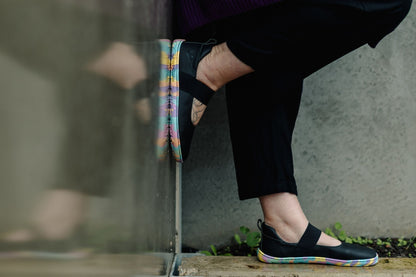 A close-up lifestyle shot of a person wearing black slip-on PaperKrane mary janes with a colourful, rainbow-like sole and black pants, sitting against a concrete wall.
