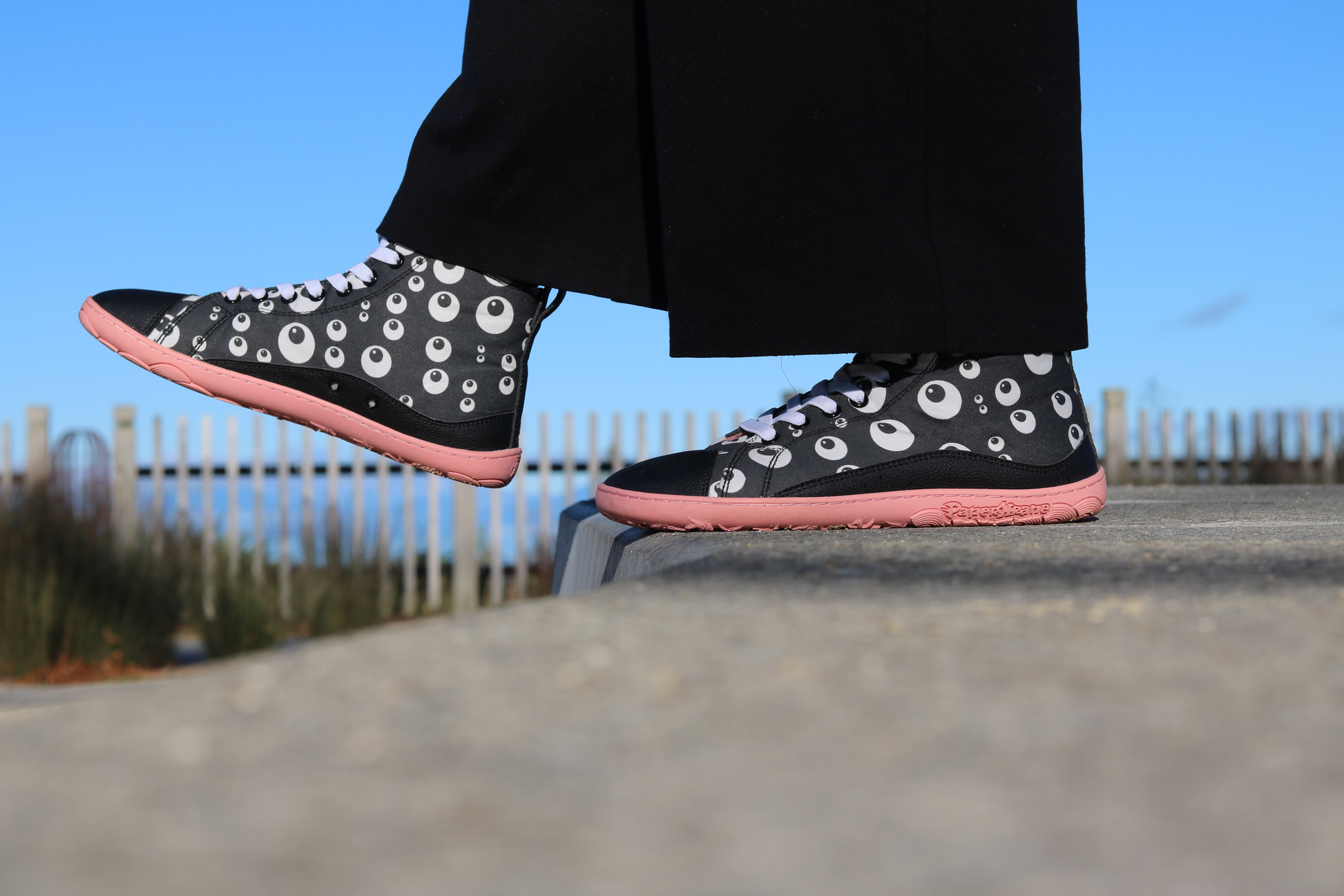 PaperKrane Eyeball boots, barefoot soles in Salmon colour, with elastic laces and black printed cotton. The print is a cartoon eyeball print, and the person is standing on a ledge and stepping off with one foot extended. The sky is blue. 