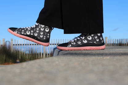 PaperKrane Eyeball boots, barefoot soles in Salmon colour, with elastic laces and black printed cotton. The print is a cartoon eyeball print, and the person is standing on a ledge and stepping off with one foot extended. The sky is blue. 