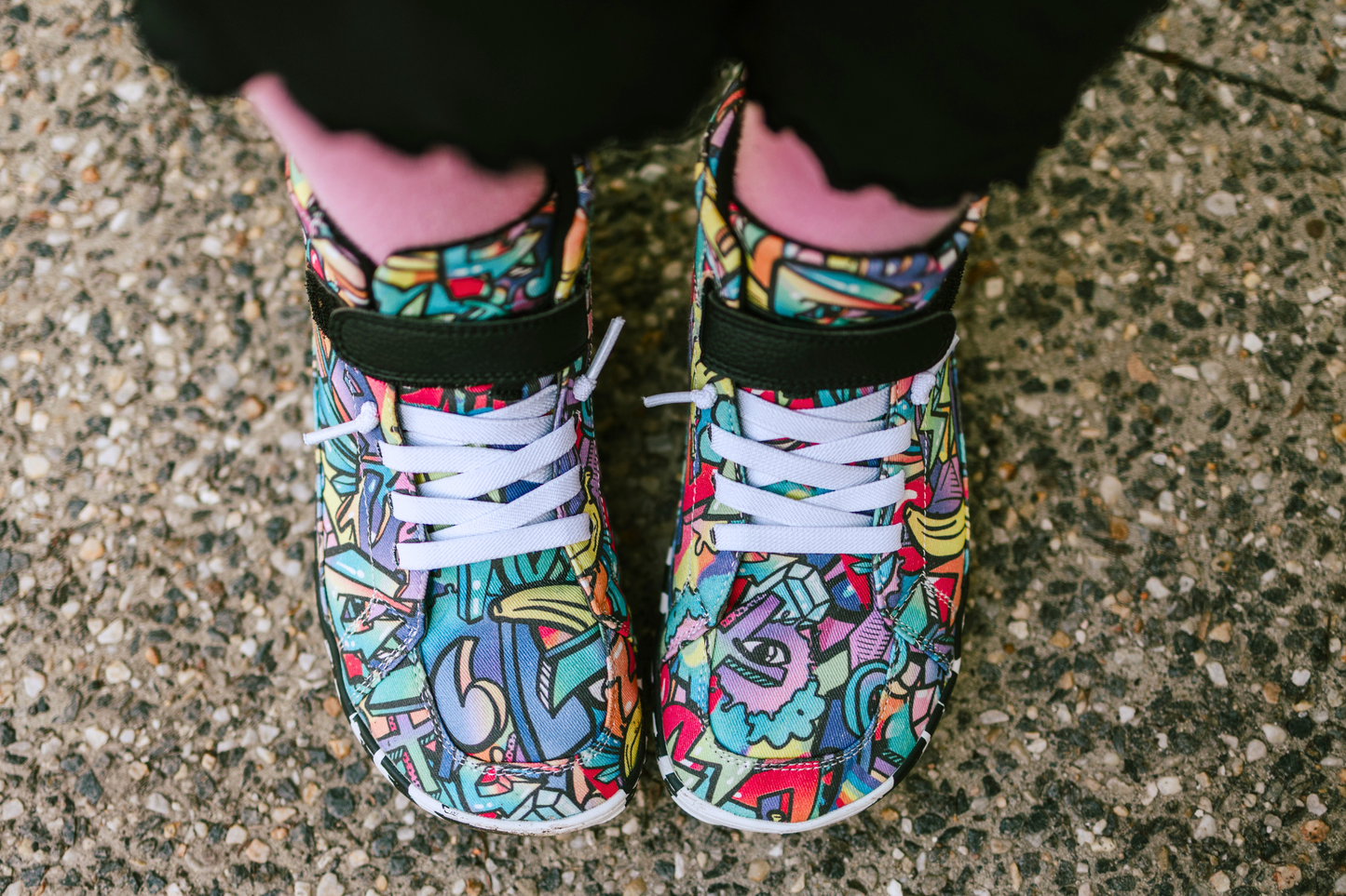 PaperKrane foot-shaped Mid Top barefoot shoes with graffiti print on cotton, black velcro strap, and zebra soles. The shoes are on a child with pink socks and black pants, the view is from above the toe box, showing their shape. 