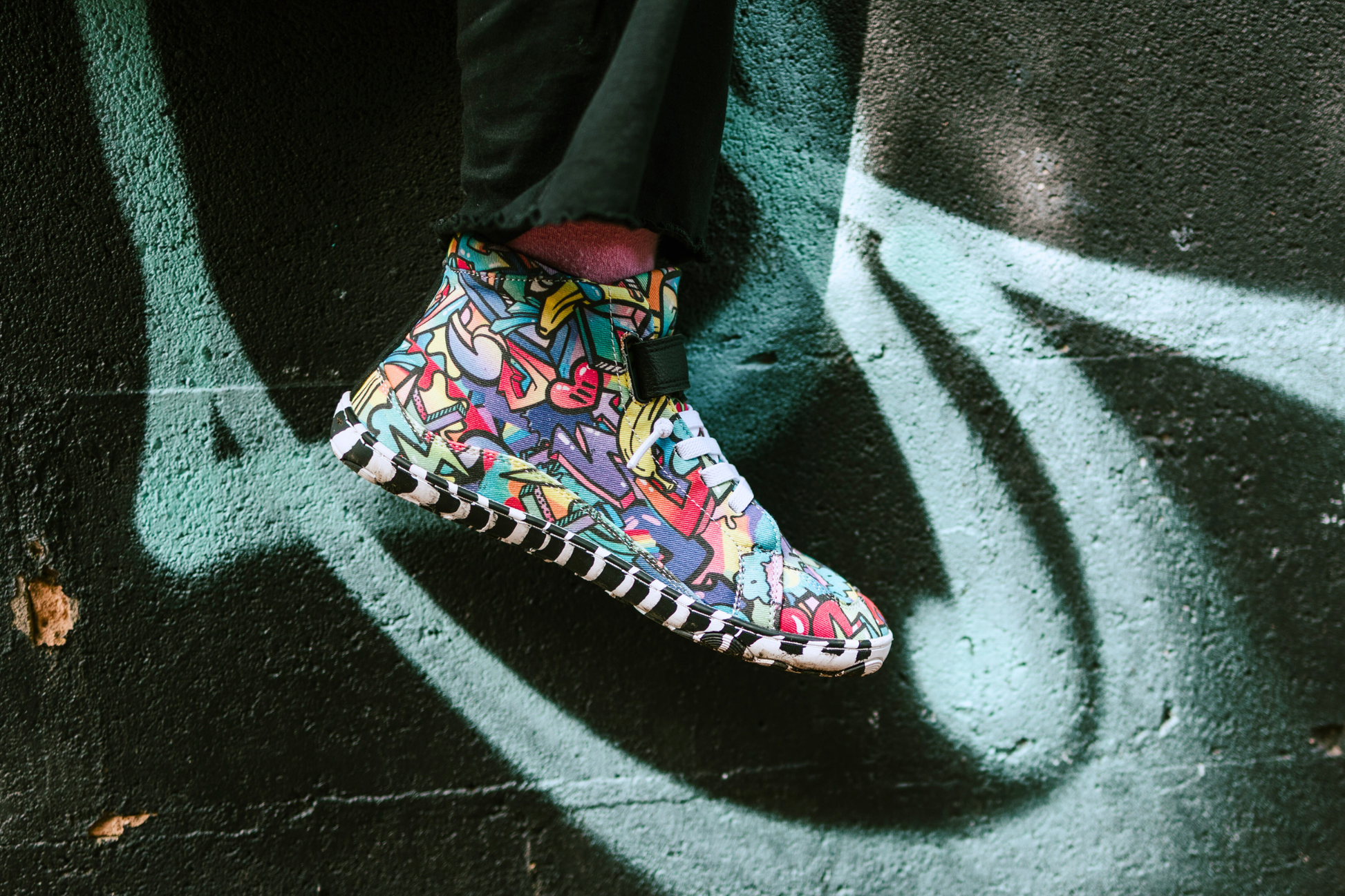 PaperKrane foot-shaped Mid Top barefoot shoes with graffiti print on cotton, black velcro strap, and zebra soles. One foot dangles in front of a brick wall with graffiti on it. The shoes are worn by a child with pink socks and black pants. 