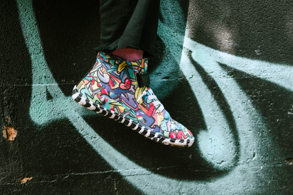 PaperKrane foot-shaped Mid Top barefoot shoes with graffiti print on cotton, black velcro strap, and zebra soles. One foot dangles in front of a brick wall with graffiti on it. The shoes are worn by a child with pink socks and black pants. 