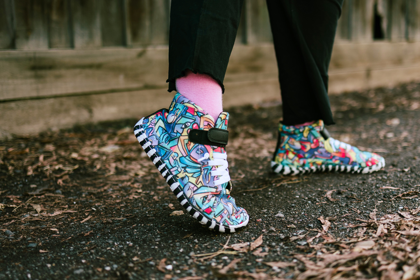 PaperKrane foot-shaped Mid Top barefoot shoes with graffiti print on cotton, black velcro strap, and zebra soles. A child with pink socks and black pants is standing on some gravel and leaves, with the back foot stretched back. 