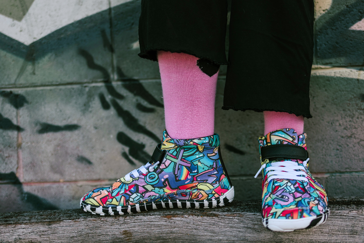PaperKrane foot-shaped Mid Top barefoot shoes with graffiti print on cotton, black velcro strap, and zebra soles. Worn by a child on a wooden surface in front of a brick wall. The child has pink socks on. 