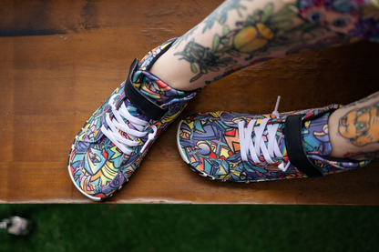 PaperKrane foot-shaped Mid Top barefoot shoes with graffiti print on cotton, black velcro strap, and zebra soles. The shoes can be seen from above on an adult's tattooed legs. They're sitting on a brown wooden bench. 