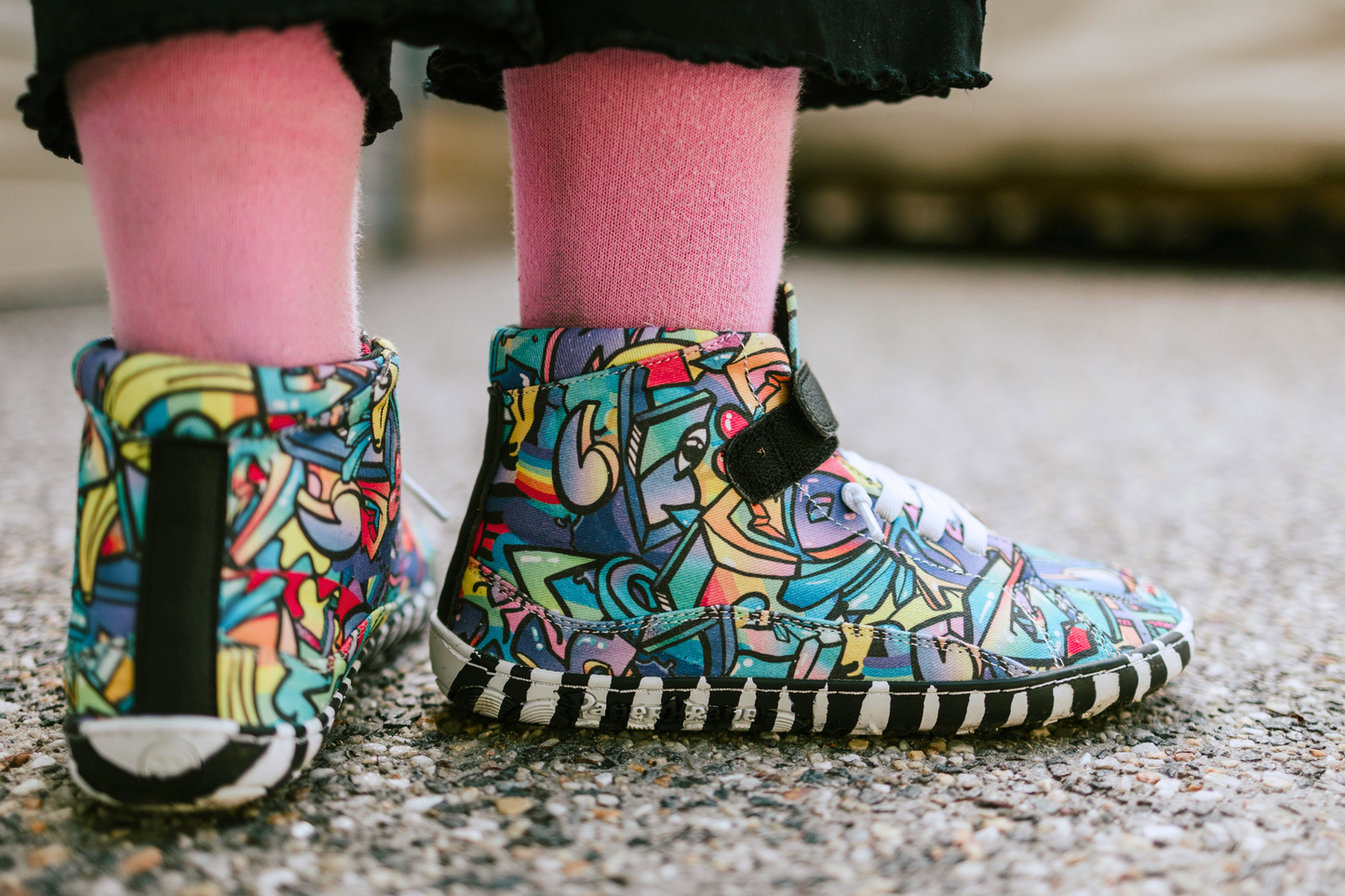 PaperKrane foot-shaped Mid Top barefoot shoes with graffiti print on cotton, black velcro strap, and zebra soles. A child with pink socks wears the Graffiti Midtops on pavement. Showing the back and side of their shoes. 