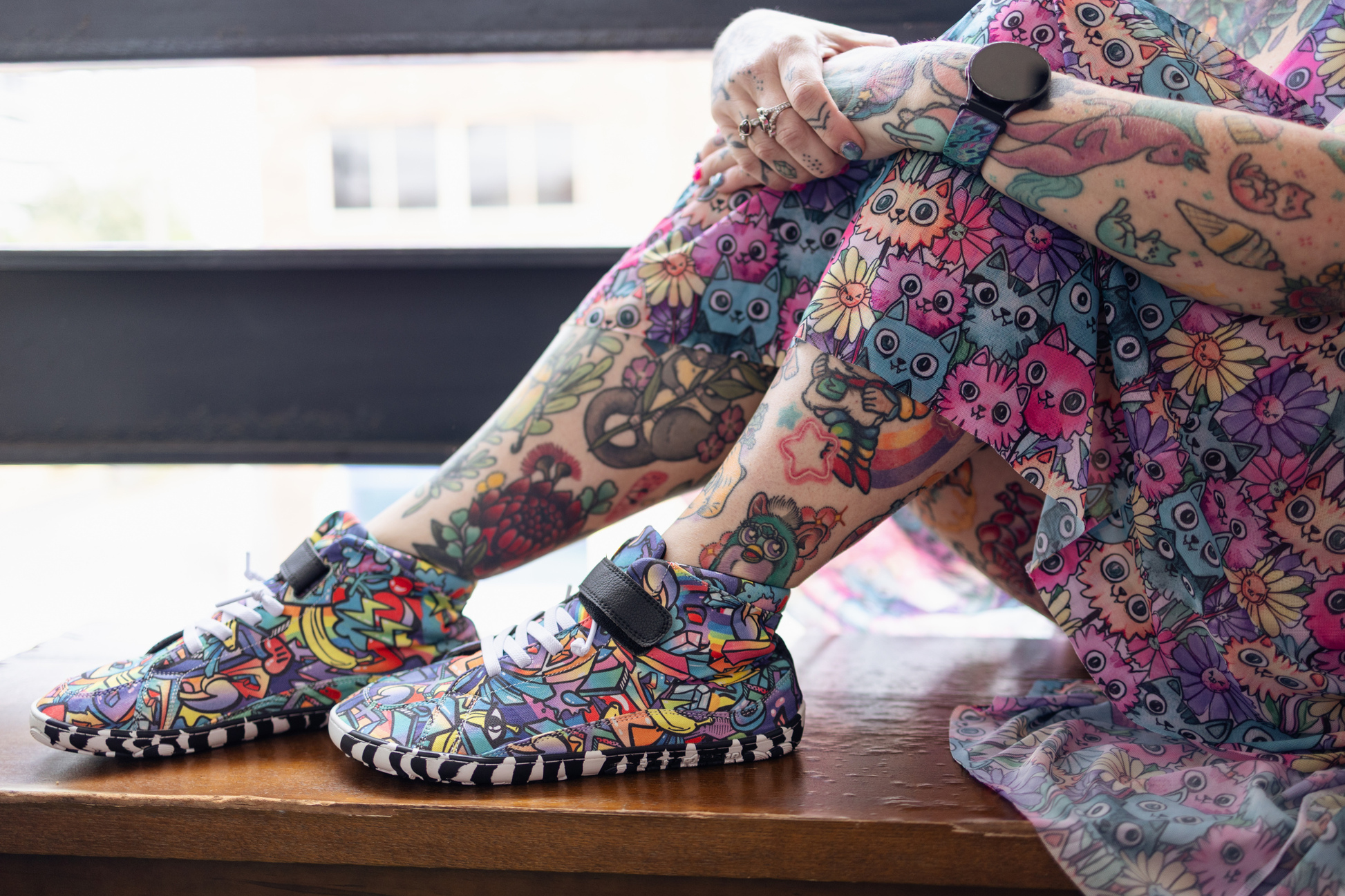 PaperKrane foot-shaped Mid Top barefoot shoes with graffiti print on cotton, black velcro strap, and zebra soles. Adult with tattoos, and colourful cat dress, is sitting down with the graffiti shoes in front of them on a bench. 
