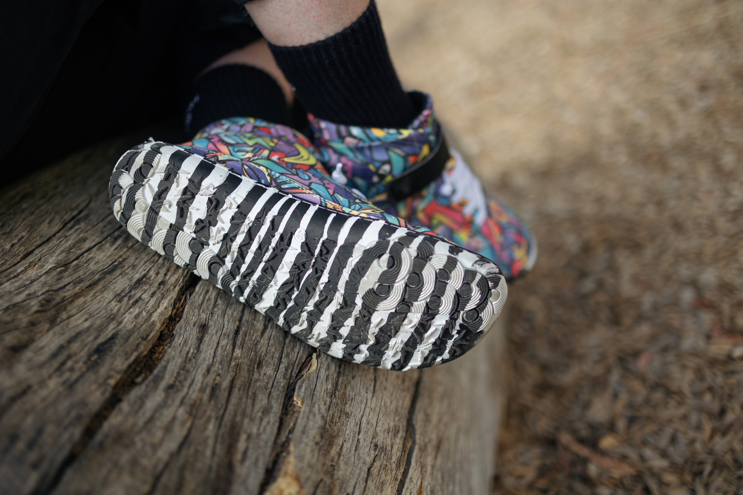 PaperKrane foot-shaped Mid Top barefoot shoes with graffiti print on cotton, black velcro strap, and zebra soles. The person is sitting on a log and showing one sole of their Graffiti mid tops. The sole is zebra style black and white stripes. 