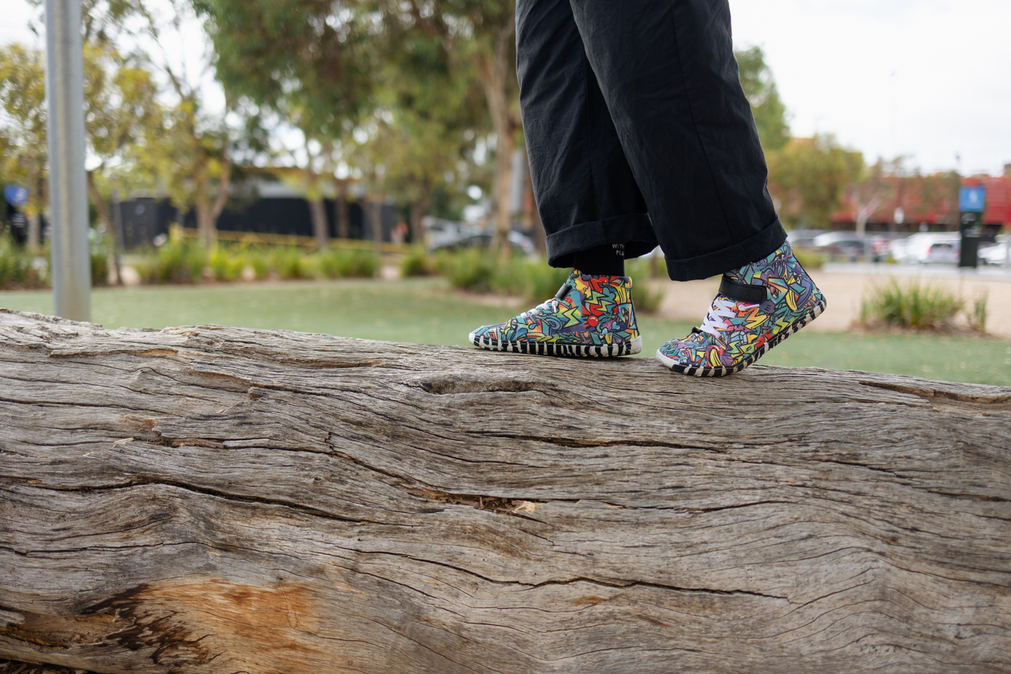 PaperKrane foot-shaped Mid Top barefoot shoes with graffiti print on cotton, black velcro strap, and zebra soles. An adult in black pants is walking on top of a log to show how balanced you are in barefoot shoes, and to show off the fun graffiti print on the shoes. 