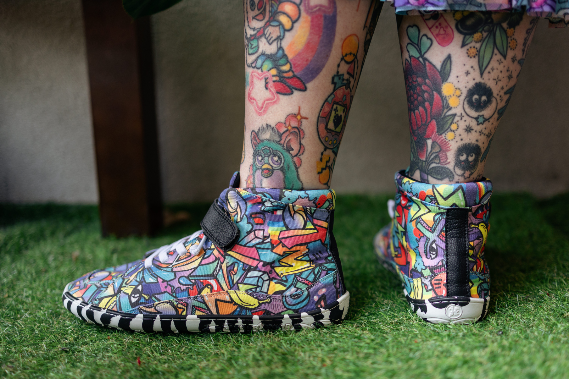 PaperKrane foot-shaped Mid Top barefoot shoes with graffiti print on cotton, black velcro strap, and zebra soles. On tattooed adult legs, on artifical grass surface.