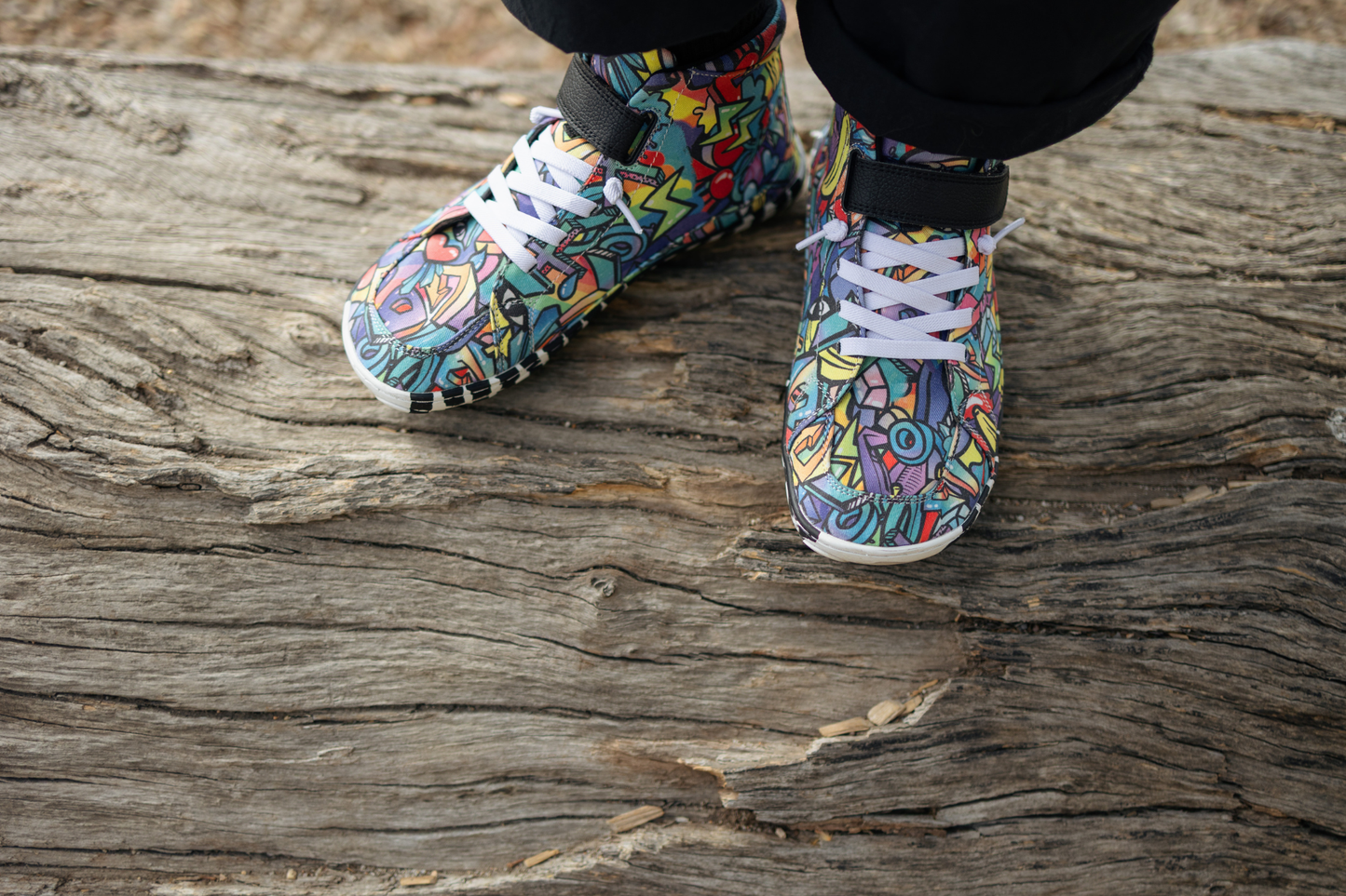 PaperKrane foot-shaped Mid Top barefoot shoes with graffiti print on cotton, black velcro strap, and zebra soles. On an adult with black pants, standing on a log. View is from an above angle.