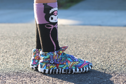 PaperKrane Graffiti signature style barefoot shoe slip ons, with hand-drawn graffiti print and zebra soles, seen on a child wearing character socks, standing on the pavement. 