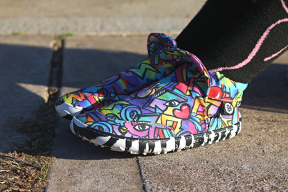 PaperKrane Graffiti signature style barefoot shoe slip ons, with hand-drawn graffiti print and zebra soles, on the feet of a child who is sitting on the footath. The shoe has a pop up tongue, and elastic ankles. 