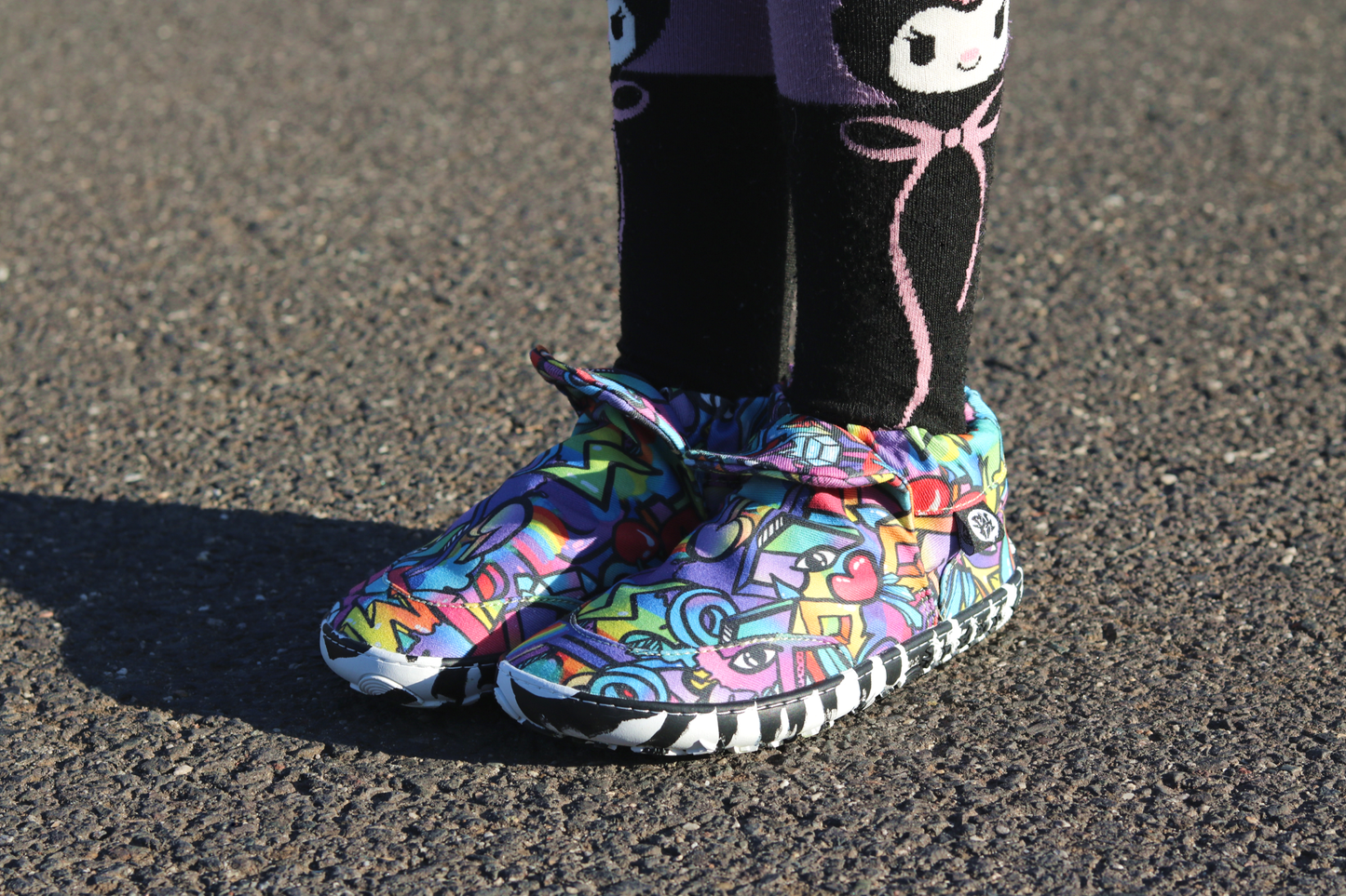 PaperKrane Graffiti signature style barefoot shoe slip ons, with hand-drawn graffiti print and zebra soles, on a child who is standing on pavement, also wearing character socks.