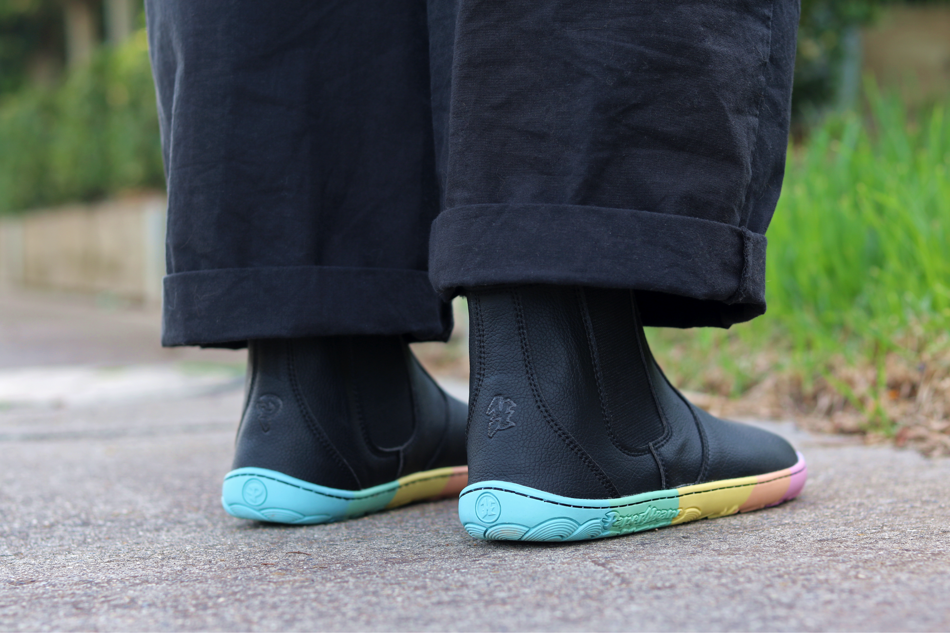 Back view of black vegan chelsea boots with a zero drop rainbow sole from PaperKrane. A person wearing black pants with folded cuffs is showing the back of the boots while standing on a footpath in front of some grass. 